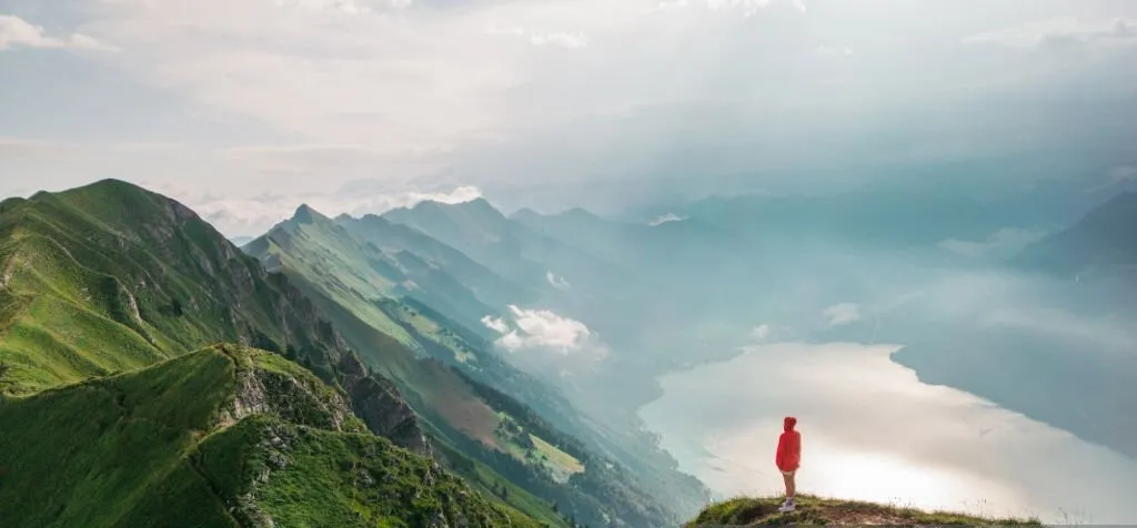 A imagem, tirada de uma perspectiva aérea em um dia claro, mostra uma pessoa vestindo uma jaqueta vermelha em pé na borda de um cume de montanha, olhando para um lago e uma vasta paisagem montanhosa.
Primeiro Plano: A encosta íngreme do cume, coberta por grama verde, leva o olhar até a pessoa em primeiro plano, no canto inferior direito.
Plano de Fundo: A paisagem se abre para um grande corpo de água espelhada abaixo e, além, múltiplas camadas de picos de montanhas que se estendem até o horizonte. Uma névoa suave envolve os vales distantes.
Céu: O céu é claro, com algumas nuvens brancas esparsas.
A cena evoca um uma sensação de aventura, isolamento e escala da natureza.